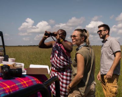 African American tour guide looking through binoculars while standing next to his tourists during lunch break by the jeep in nature reserve.
