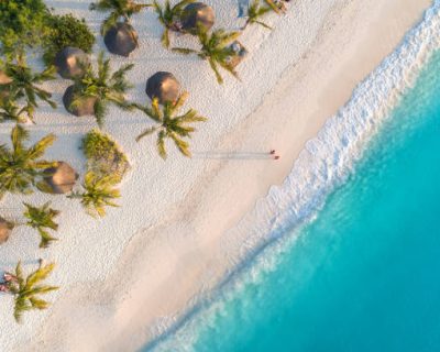 Aerial view of umbrellas, palms on the sandy beach of Indian Ocean at sunset. Summer holiday in Zanzibar, Africa. Tropical landscape with palm trees, parasols, white sand, blue water, waves. Top view